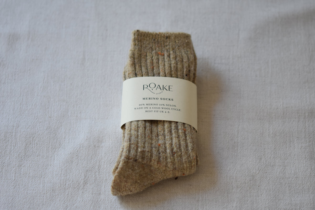A pair of beige fleck ribbed knit Merino wool socks folded and wrapped with cream paper band reading Roake Studio, on a light fabric background.