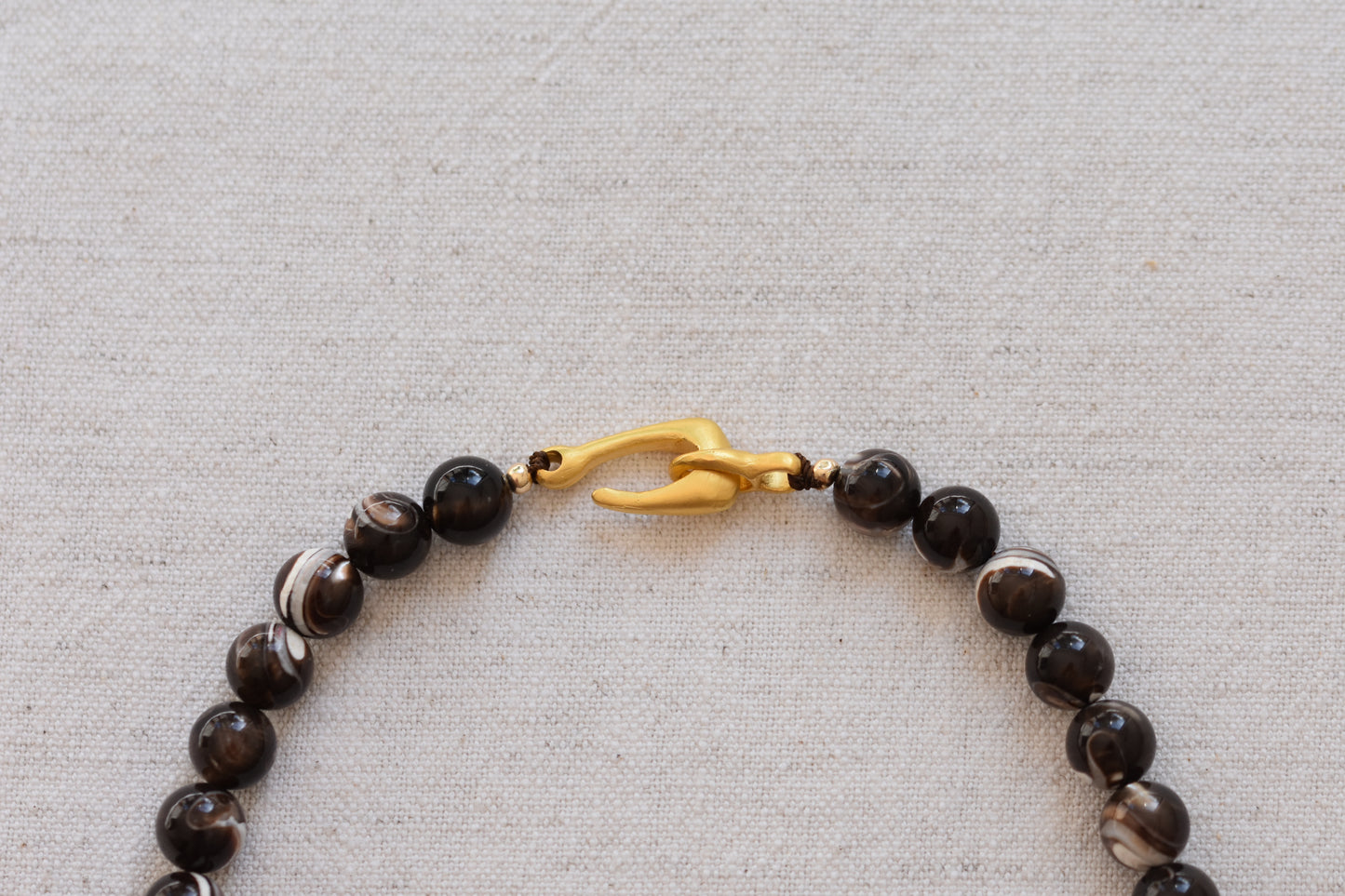 By my hands Burnt Umber Bei Necklace of opalescent dark brown Mother of Pearl beads, with a hand-carved gold vermeil fish hook closure, close up on a beige background.
