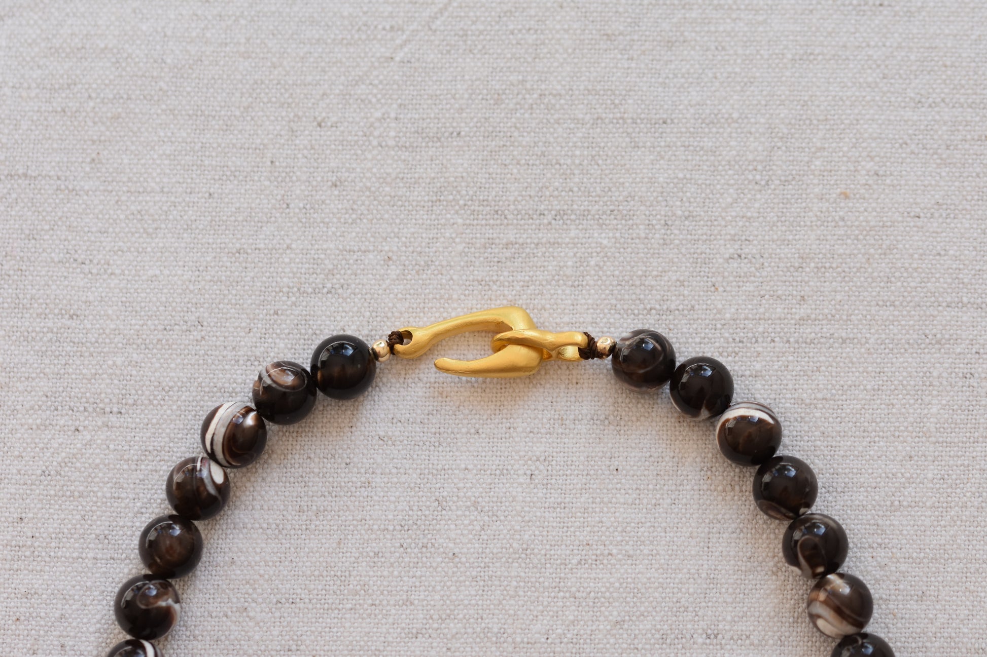 By my hands Burnt Umber Bei Necklace of opalescent dark brown Mother of Pearl beads, with a hand-carved gold vermeil fish hook closure, close up on a beige background.