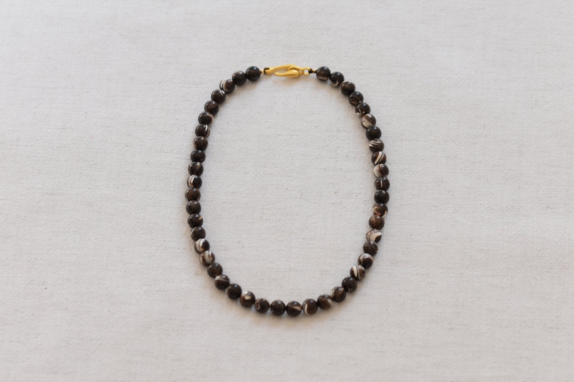 By my hands Burnt Umber Bei Necklace of opalescent dark brown Mother of Pearl beads, with a hand-carved gold vermeil fish hook closure, on a beige background.