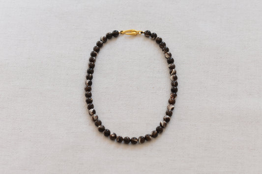 By my hands Burnt Umber Bei Necklace of opalescent dark brown Mother of Pearl beads, with a hand-carved gold vermeil fish hook closure, on a beige background.