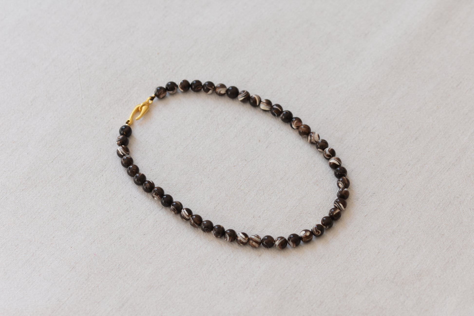 By my hands Burnt Umber Bei Necklace of opalescent dark brown Mother of Pearl beads, with a hand-carved gold vermeil fish hook closure, on a beige background.