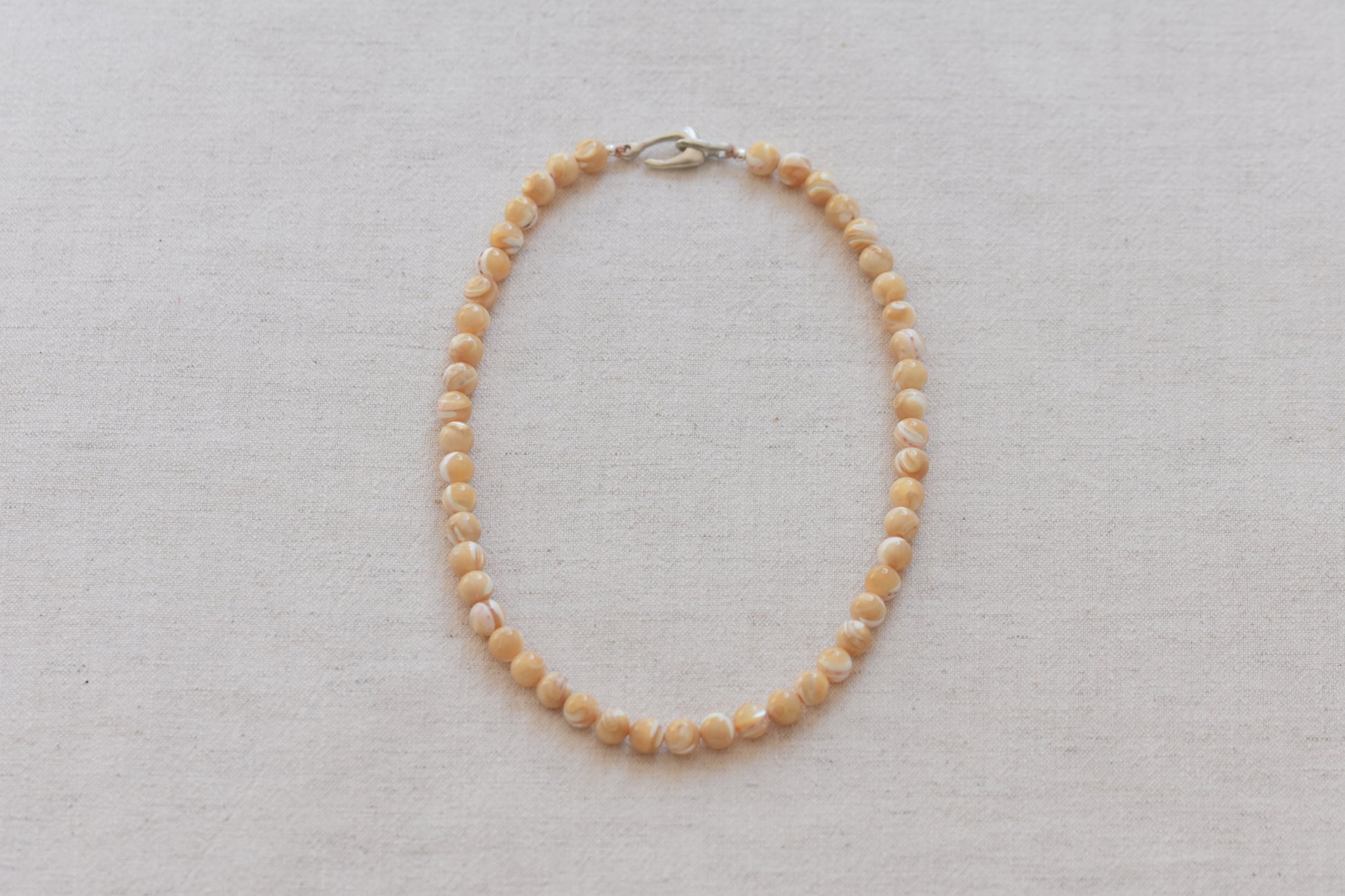 By my hands Sandy Bei Necklace of opalescent light yellow Mother of Pearl beads, with a hand-carved sterling silver fish hook closure, on a beige background.