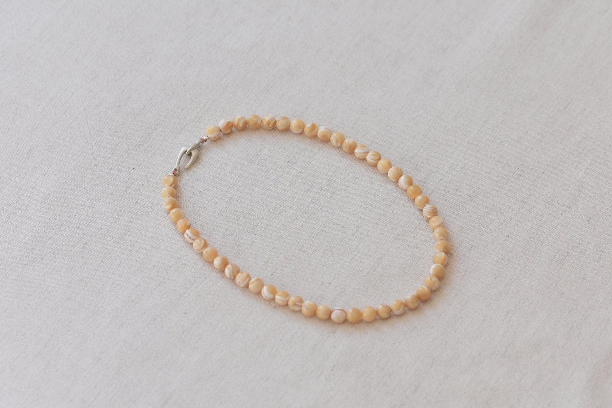 By my hands Sandy Bei Necklace of opalescent light yellow Mother of Pearl beads, with a hand-carved sterling silver fish hook closure, on a beige background.