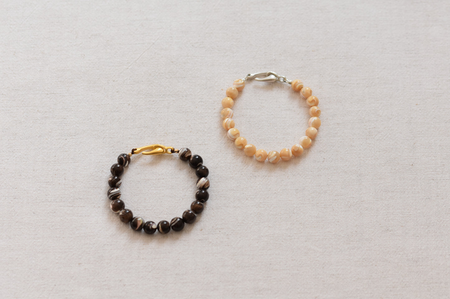 By my hands Burnt Umber Bei Bracelet of dark brown and Sandy Bei Bracelet of light yellow Mother of Pearl beads, on a beige background.