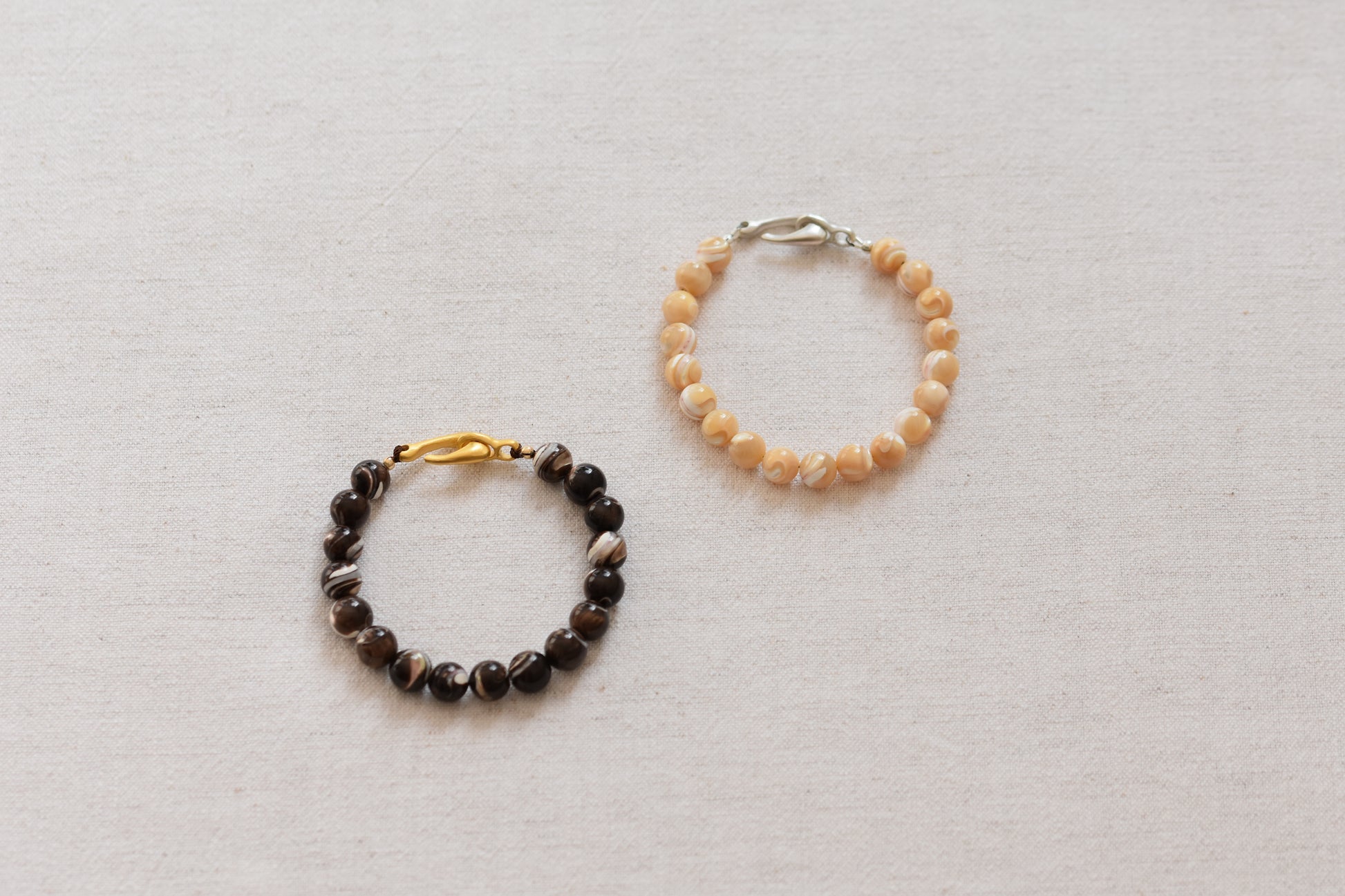 By my hands Burnt Umber Bei Bracelet of dark brown and Sandy Bei Bracelet of light yellow Mother of Pearl beads, on a beige background.