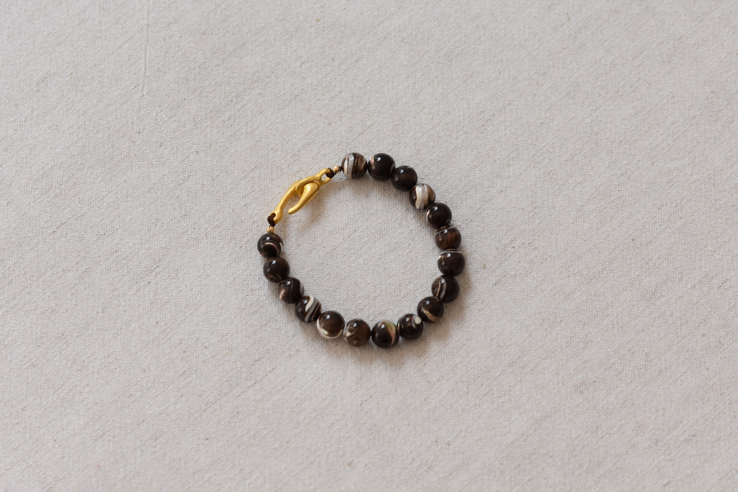 By my hands Burnt Umber Bei Bracelet of opalescent dark brown Mother of Pearl beads, with a hand-carved gold vermeil fish hook closure, on a beige background.