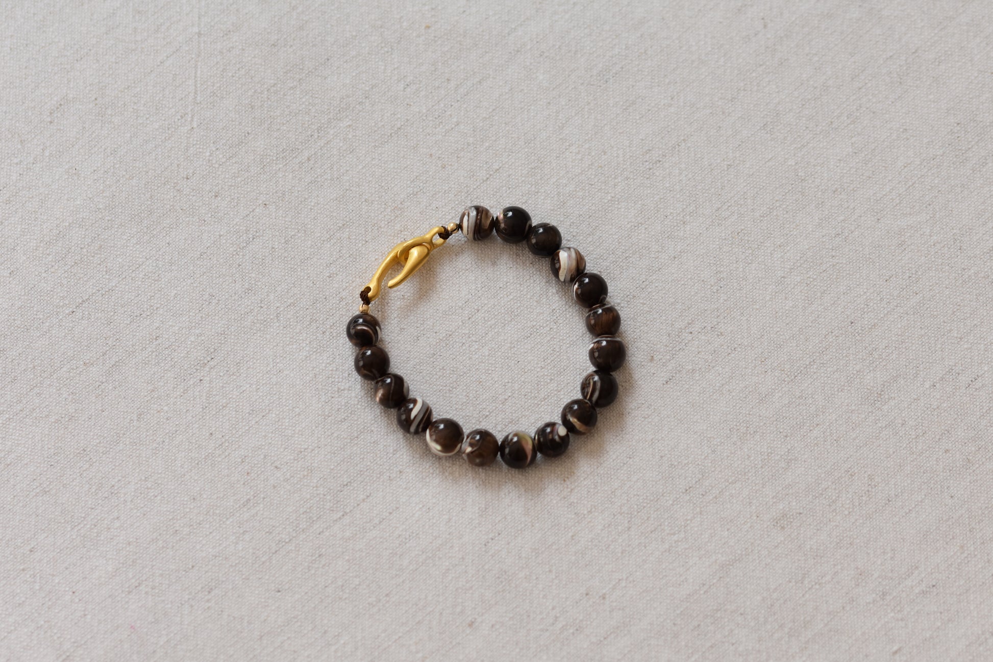 By my hands Burnt Umber Bei Bracelet of opalescent dark brown Mother of Pearl beads, with a hand-carved gold vermeil fish hook closure, on a beige background.