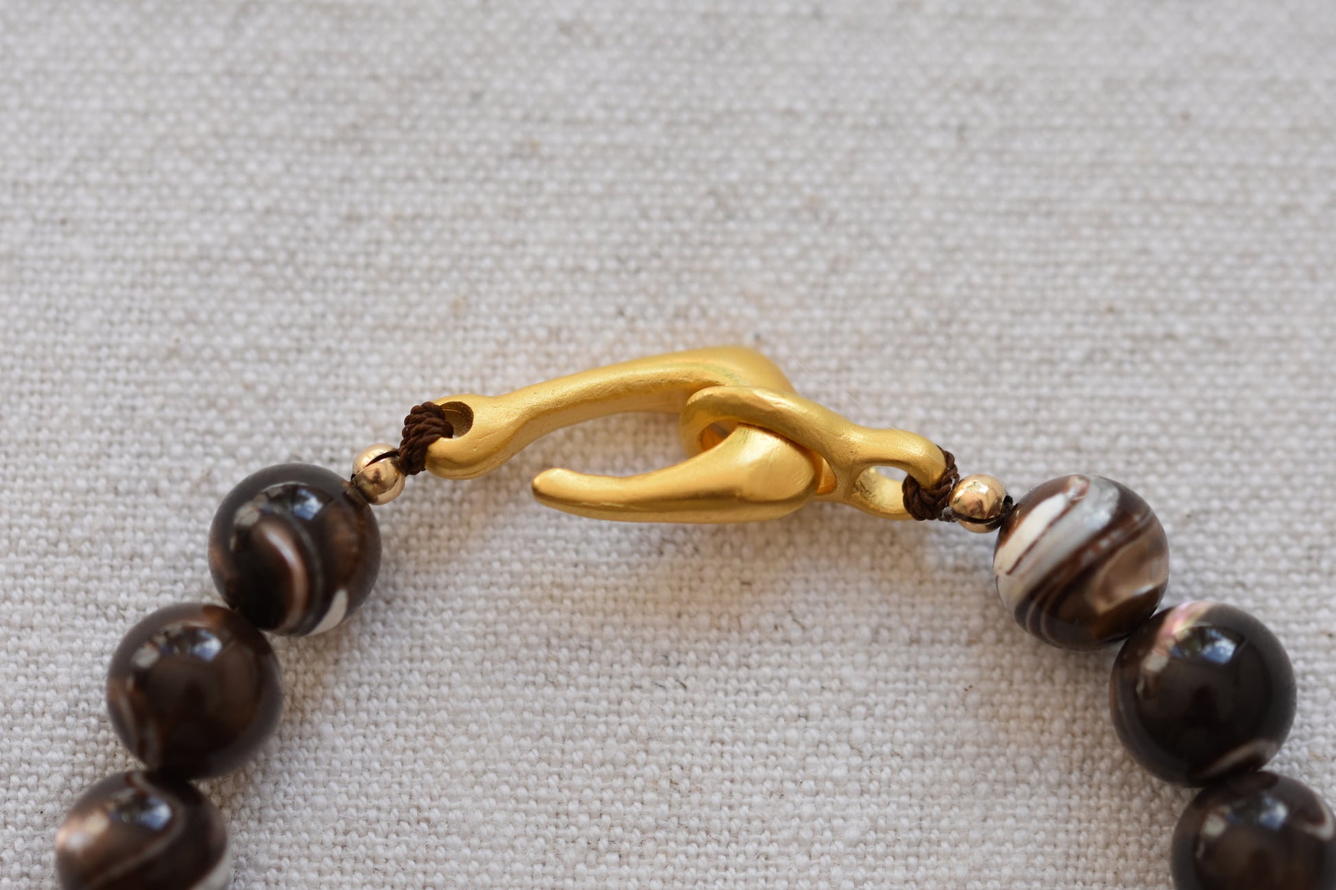 By my hands Burnt Umber Bei Bracelet of opalescent dark brown Mother of Pearl beads, with a hand-carved gold vermeil fish hook closure, close up on a beige background.