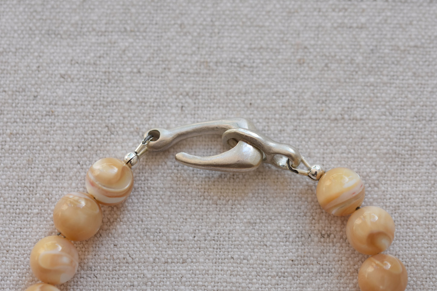 By my hands Sandy Bei Bracelet of opalescent light yellow Mother of Pearl beads, with a hand-carved sterling silver fish hook closure, close up on a beige background.