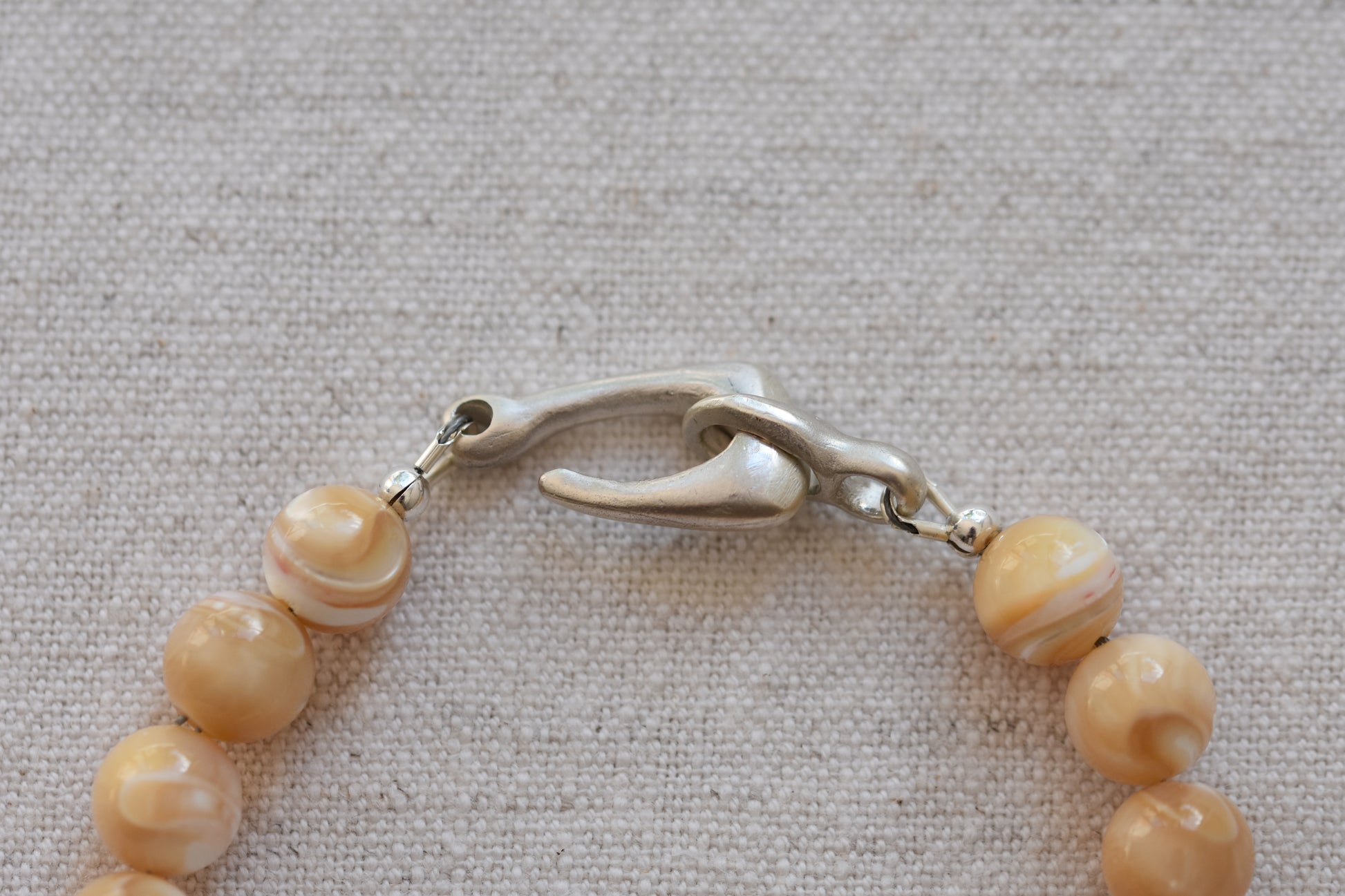 By my hands Sandy Bei Bracelet of opalescent light yellow Mother of Pearl beads, with a hand-carved sterling silver fish hook closure, close up on a beige background.