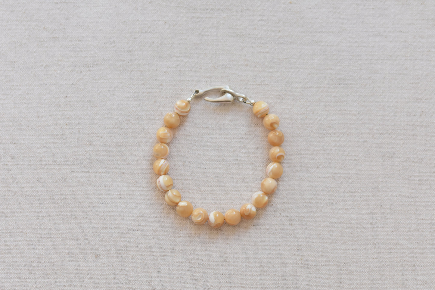 By my hands Sandy Bei Bracelet of opalescent light yellow Mother of Pearl beads, with a hand-carved sterling silver fish hook closure, on a beige background.