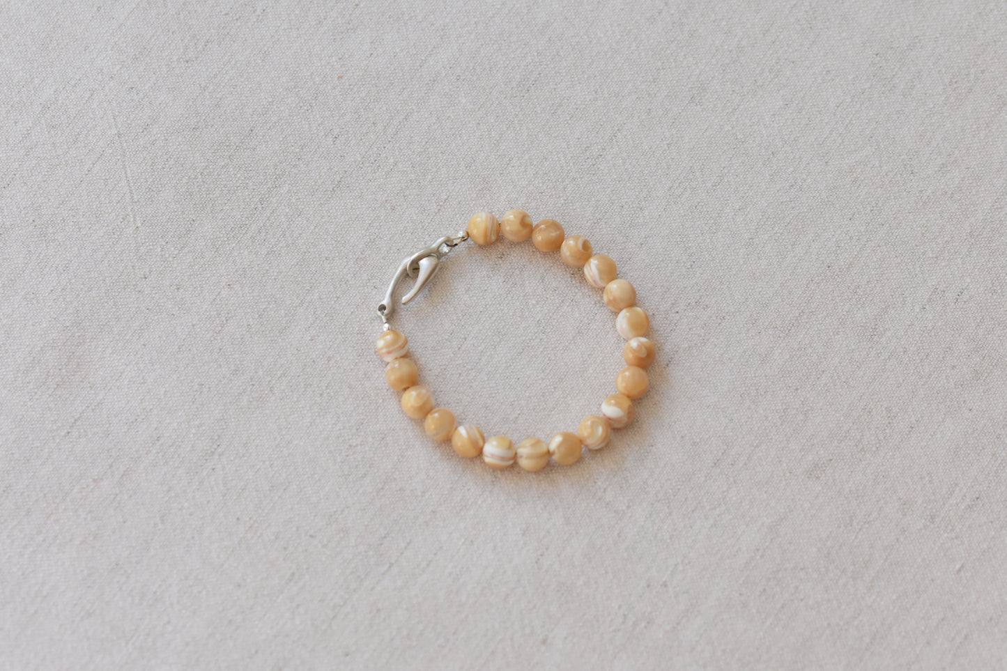By my hands Sandy Bei Bracelet of opalescent light yellow Mother of Pearl beads, with a hand-carved sterling silver fish hook closure, on a beige background.