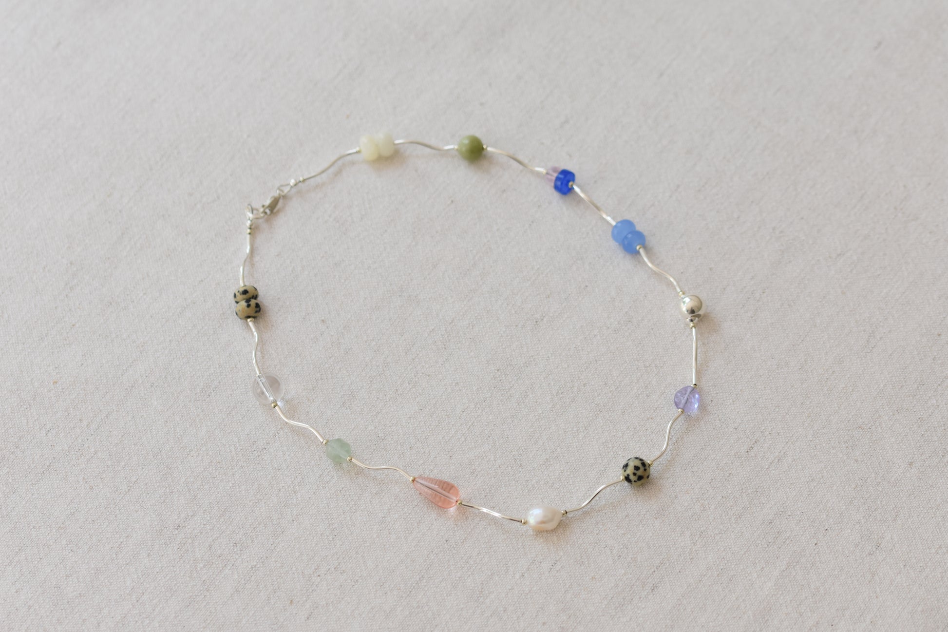 By my hands Fluorescence Ripple Necklace with wavy sterling silver ripples and gemstone beads including peridot, dalmatian jasper, freshwater pearl, and coloured jade, on a beige background.