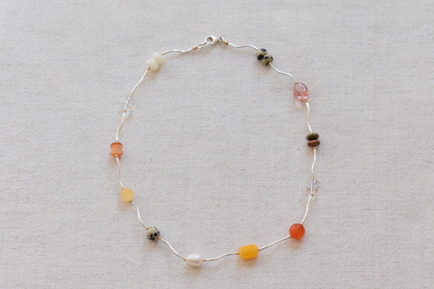 By my hands Sienna Ripple Necklace with wavy sterling silver ripples and earthy gemstones including unakite, dalmatian jasper, freshwater pearl, and coloured jade, on a beige background.