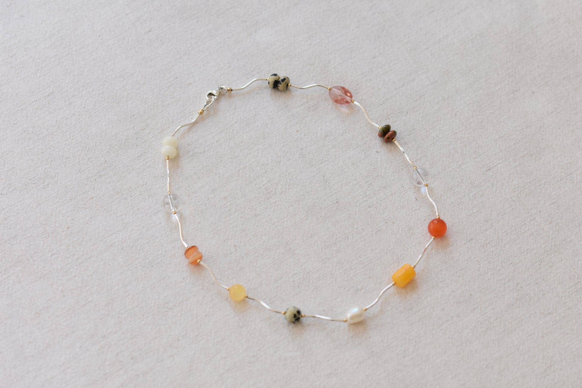 By my hands Sienna Ripple Necklace with wavy sterling silver ripples and earthy gemstones including unakite, dalmatian jasper, freshwater pearl, and coloured jade, on a beige background.