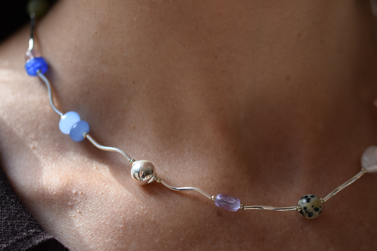 By my hands Fluorescence Ripple Necklace with wavy sterling silver ripples and gemstone beads including peridot, dalmatian jasper, freshwater pearl, and coloured jade, close up shown on a person.