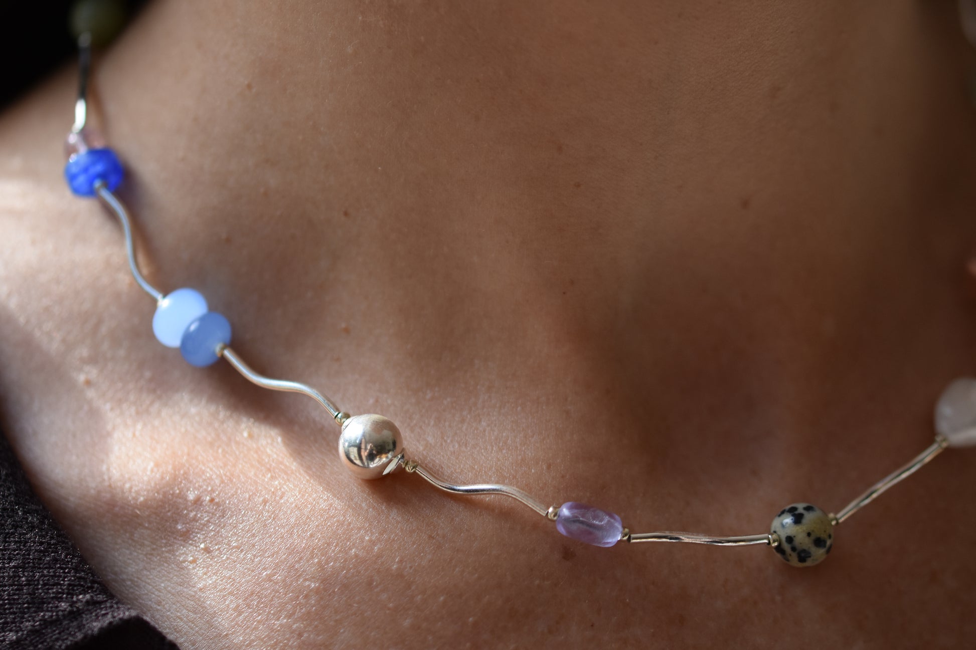 By my hands Fluorescence Ripple Necklace with wavy sterling silver ripples and gemstone beads including peridot, dalmatian jasper, freshwater pearl, and coloured jade, close up shown on a person.