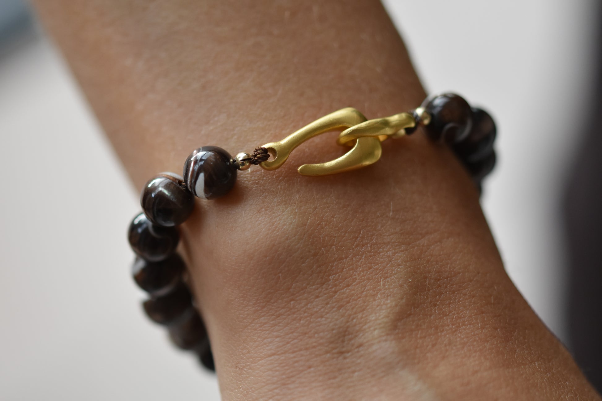 By my hands Burnt Umber Bei Bracelet of opalescent dark brown Mother of Pearl beads, with a hand-carved gold vermeil fish hook closure, shown on a person.