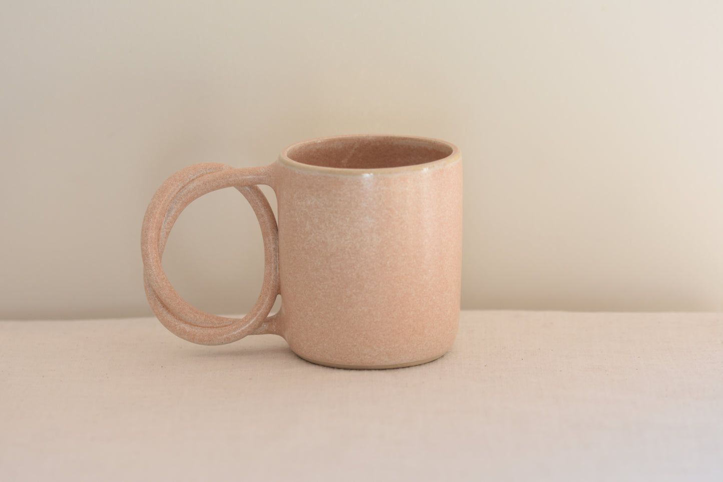 We Are Clay blush pink ceramic mug with pretzel shaped handle on a beige background.