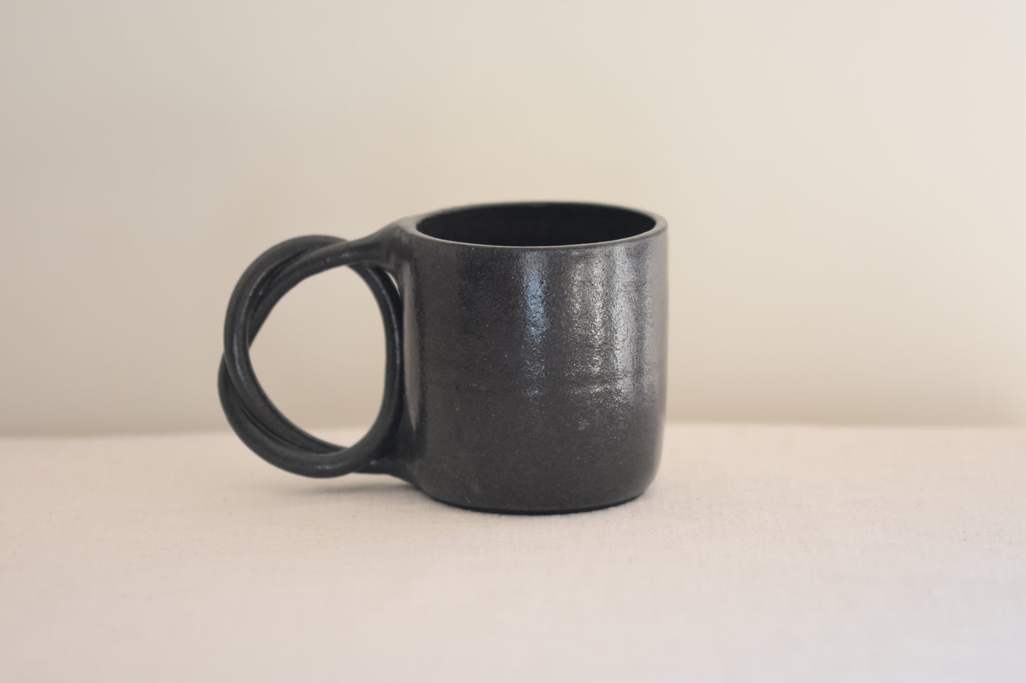We Are Clay black ceramic mug with pretzel shaped handle on a beige background.
