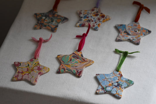 Six Jemma Lewis marbled ceramic star decorations with colourful velvet ribbons on a beige background.