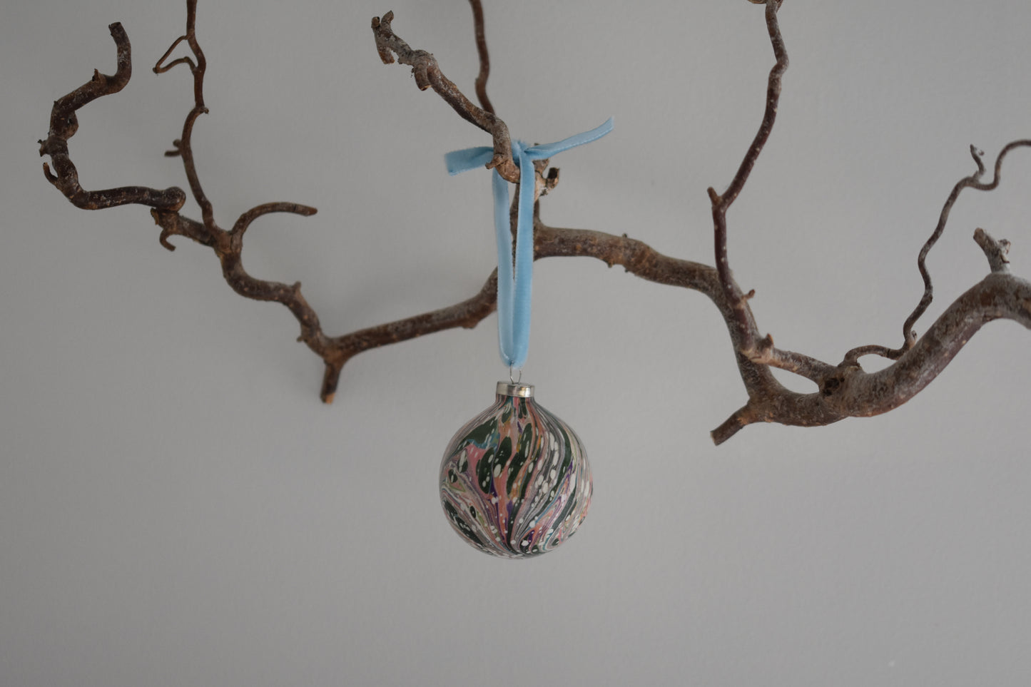 Jemma Lewis colourful marbled round ceramic bauble Christmas ornament with velvet ribbon, hanging from a branch on a white background.