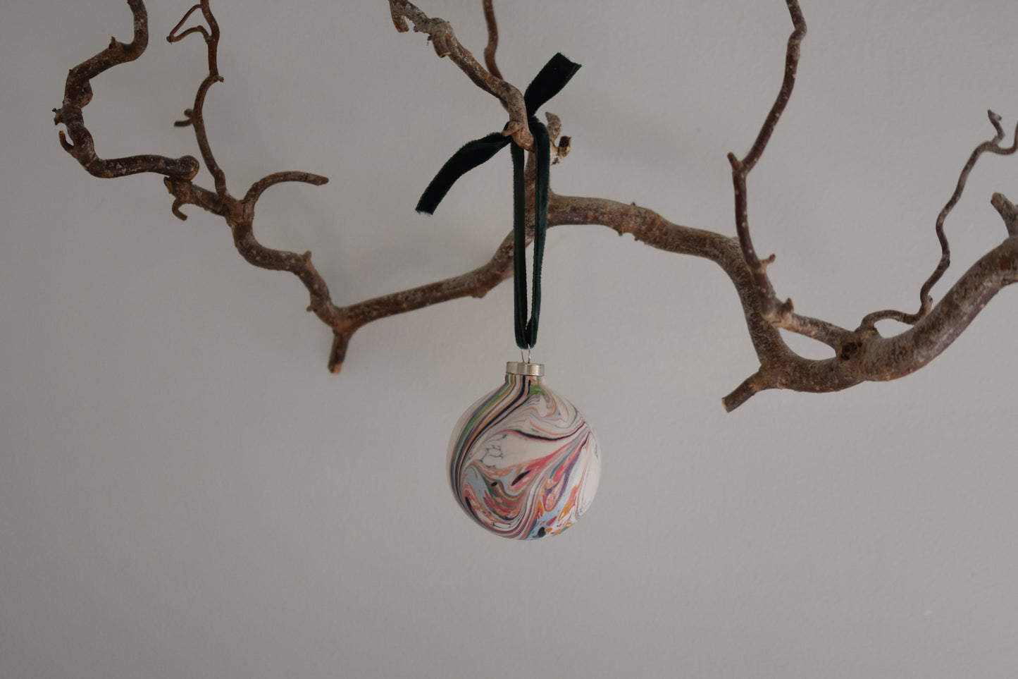 Jemma Lewis colourful marbled round ceramic bauble Christmas ornament with velvet ribbon, hanging from a branch on a white background.