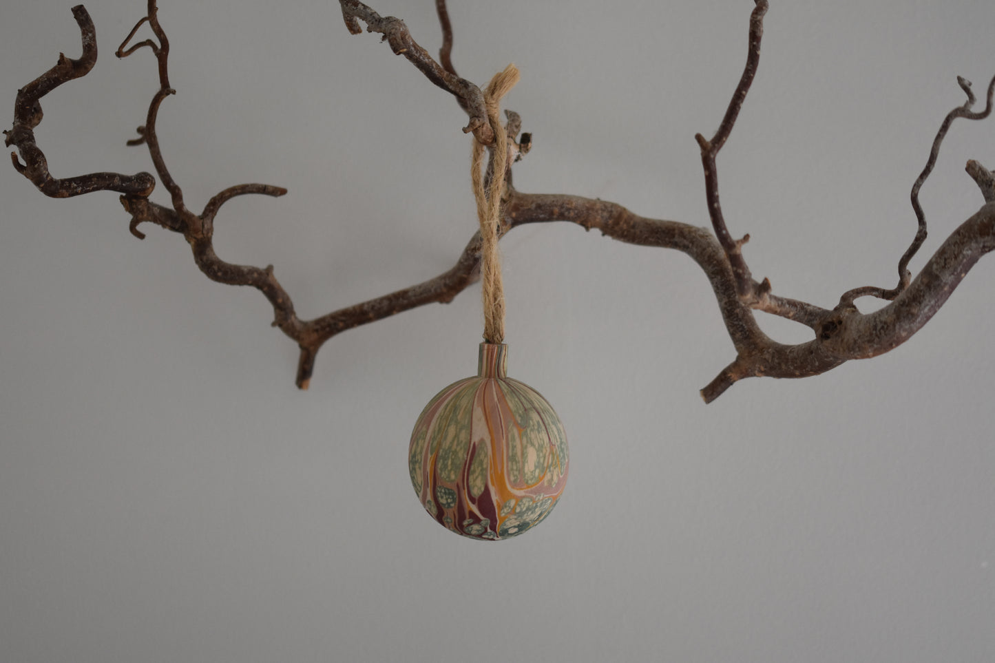 Jemma Lewis colourful marbled wooden round bauble Christmas ornament with twine, hanging from a branch on a white background.