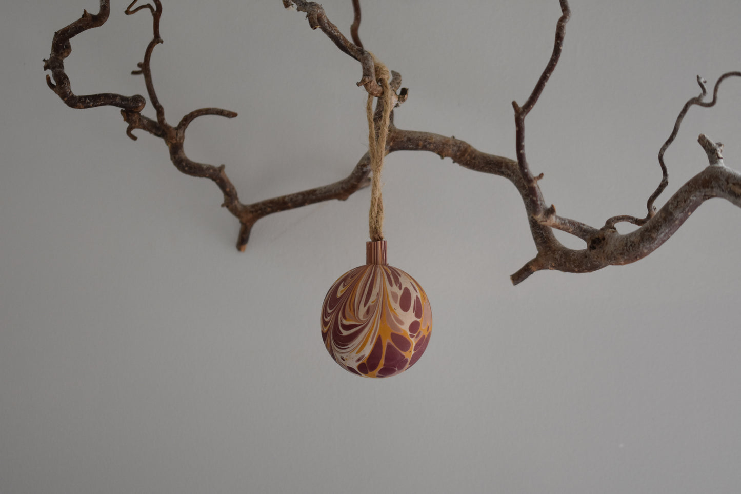 Jemma Lewis colourful marbled wooden round bauble Christmas ornament with twine, hanging from a branch on a white background.