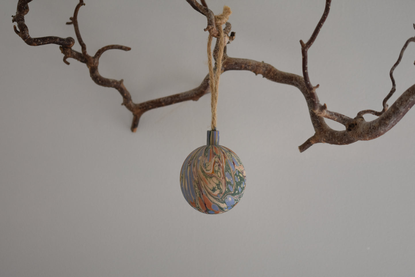 Jemma Lewis colourful marbled wooden round bauble Christmas ornament with twine, hanging from a branch on a white background.