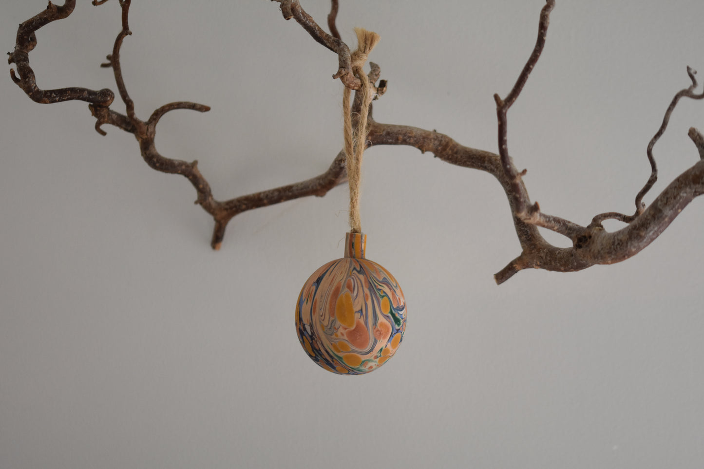 Jemma Lewis colourful marbled wooden round bauble Christmas ornament with twine, hanging from a branch on a white background.