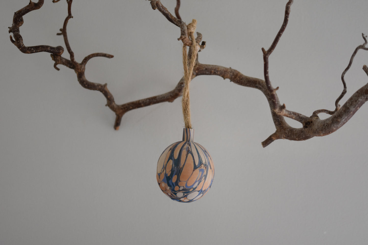 Jemma Lewis colourful marbled wooden round bauble Christmas ornament with twine, hanging from a branch on a white background.