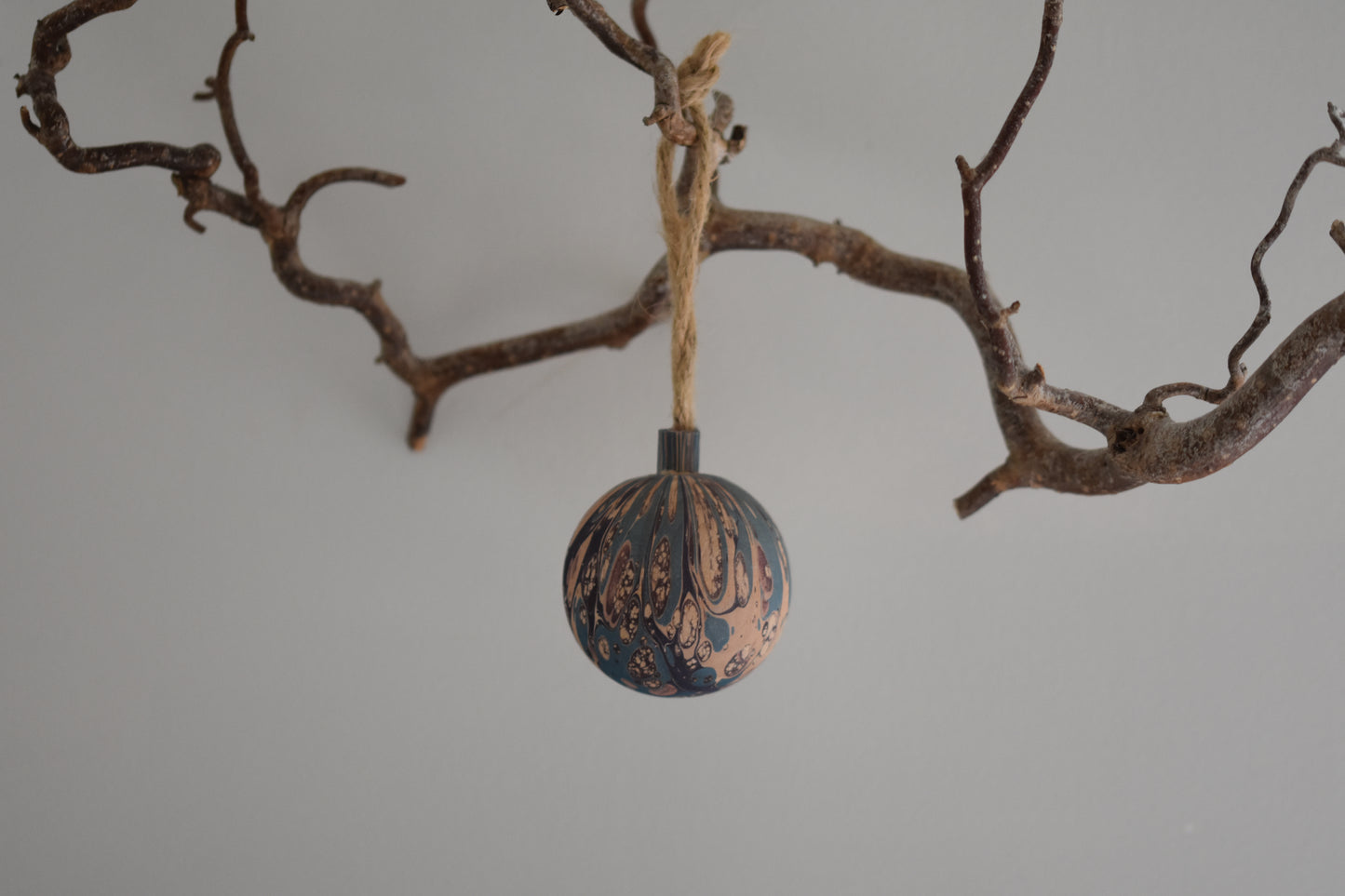 Jemma Lewis colourful marbled wooden round bauble Christmas ornament with twine, hanging from a branch on a white background.
