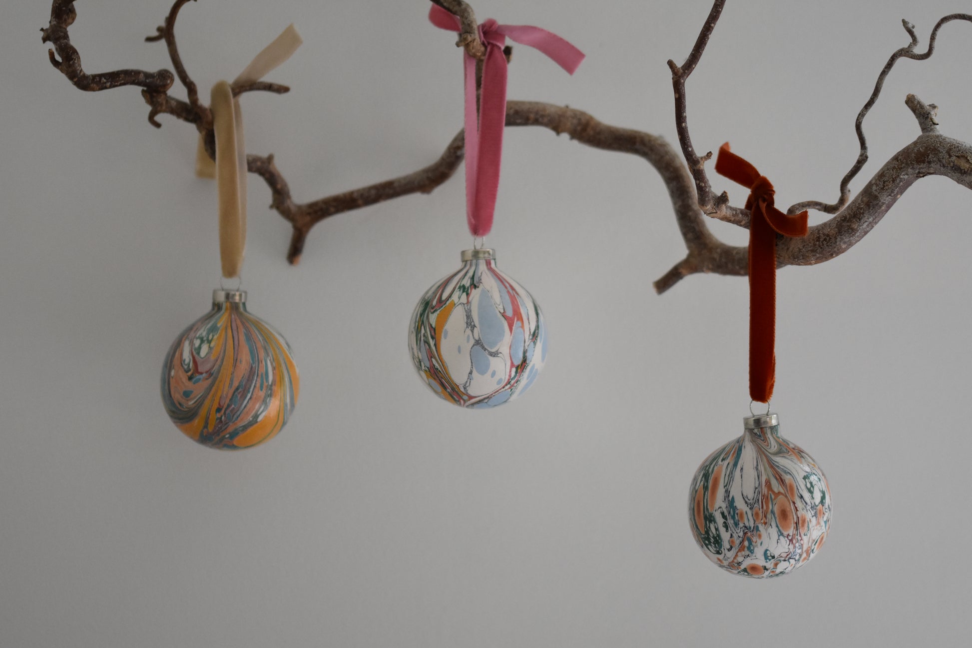 Jemma Lewis colourful marbled round ceramic bauble Christmas ornaments with velvet ribbons, hanging from a branch on a white background.