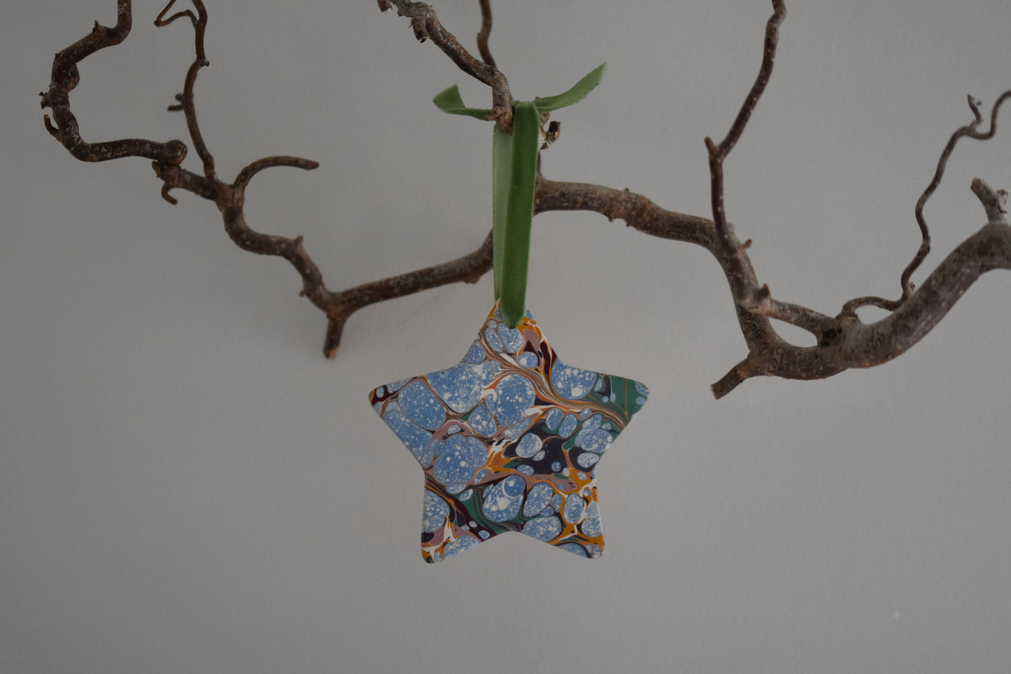 Jemma Lewis marbled ceramic star Christmas decoration ornament with colourful velvet ribbon, hanging from a branch on a white background.