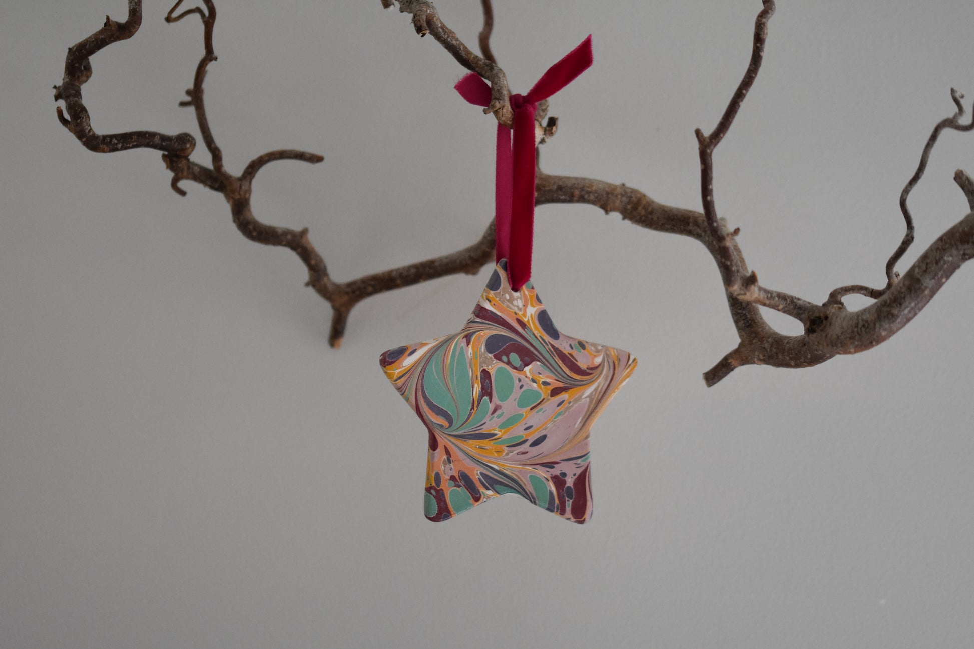 Jemma Lewis marbled ceramic star Christmas decoration ornament with colourful velvet ribbon, hanging from a branch on a white background.