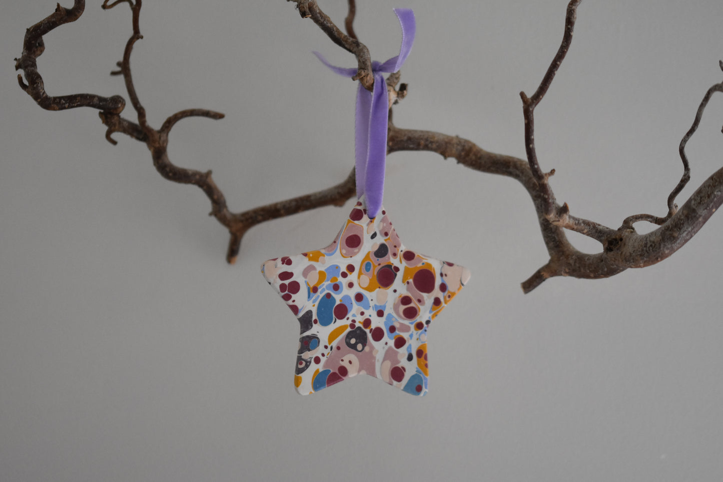 Jemma Lewis marbled ceramic star Christmas decoration ornament with colourful velvet ribbon, hanging from a branch on a white background.