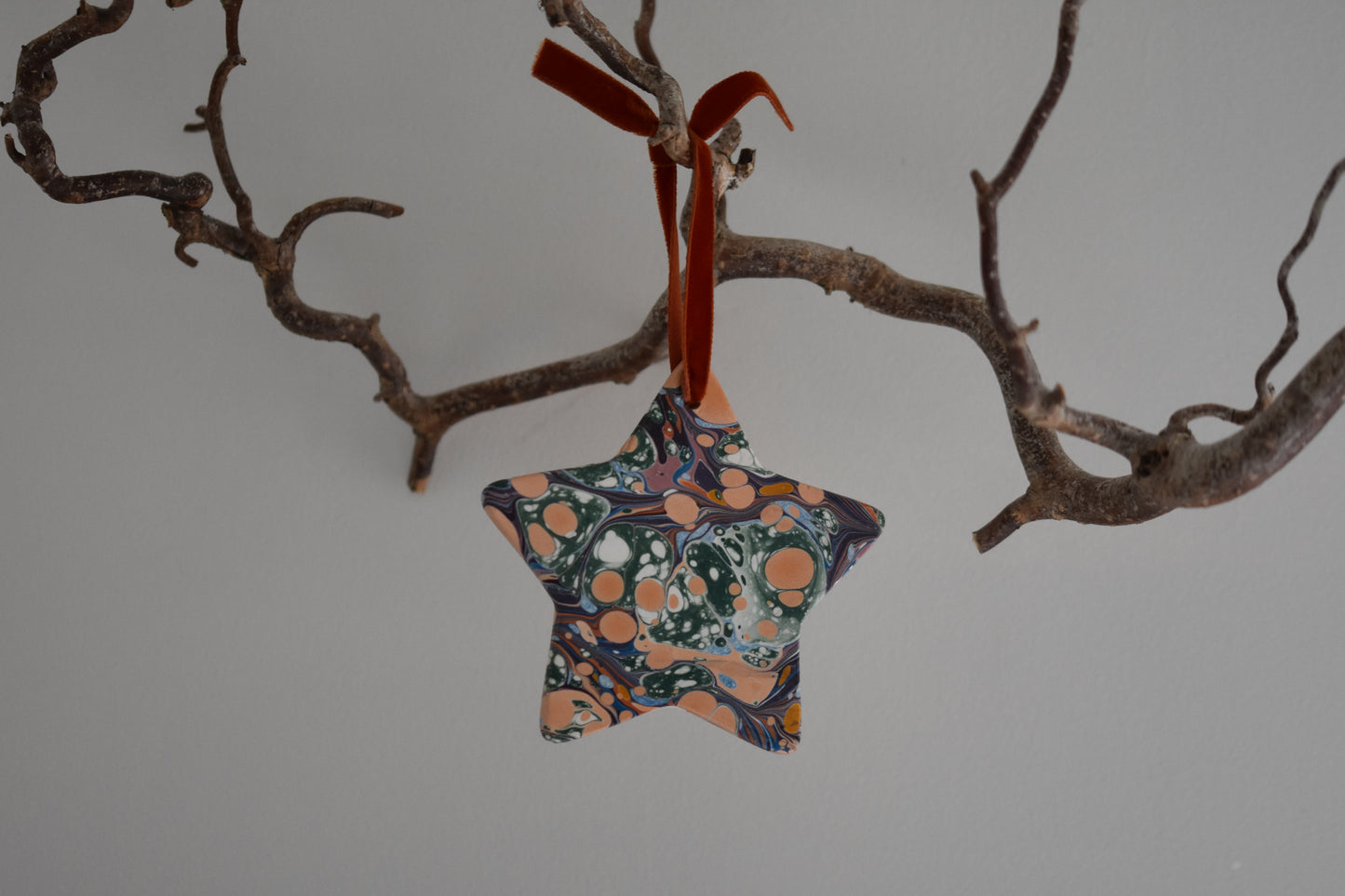Jemma Lewis marbled ceramic star Christmas decoration ornament with colourful velvet ribbon, hanging from a branch on a white background.