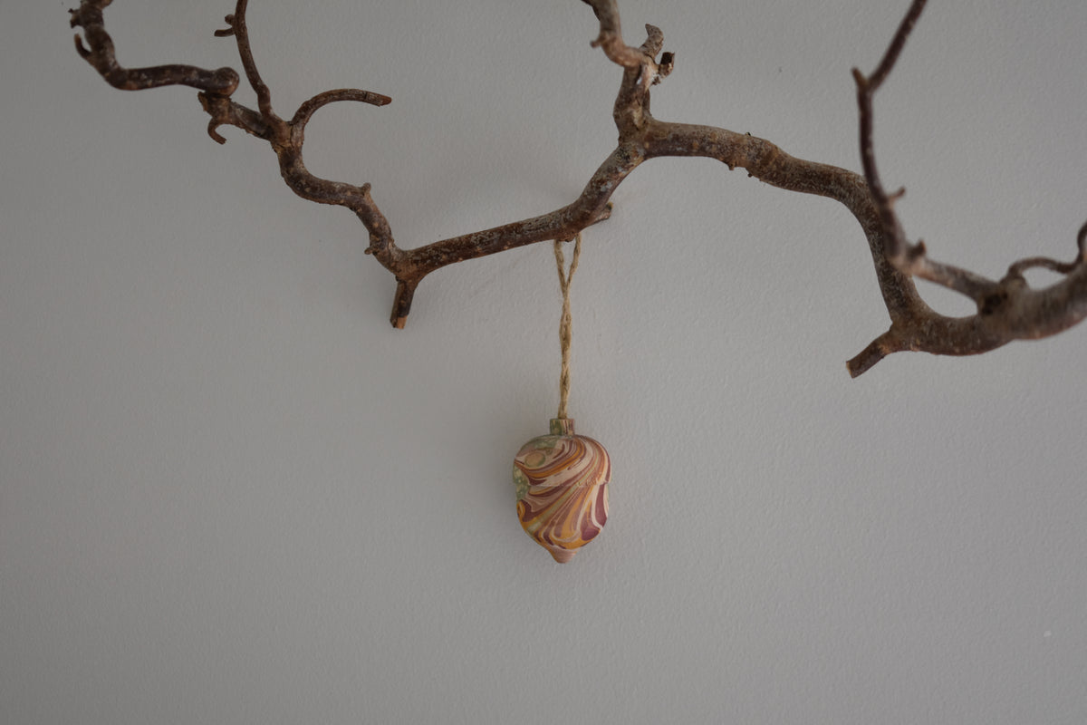 Jemma Lewis colourful marbled wooden acorn decoration Christmas ornament hung from twine on a branch, with a white background.
