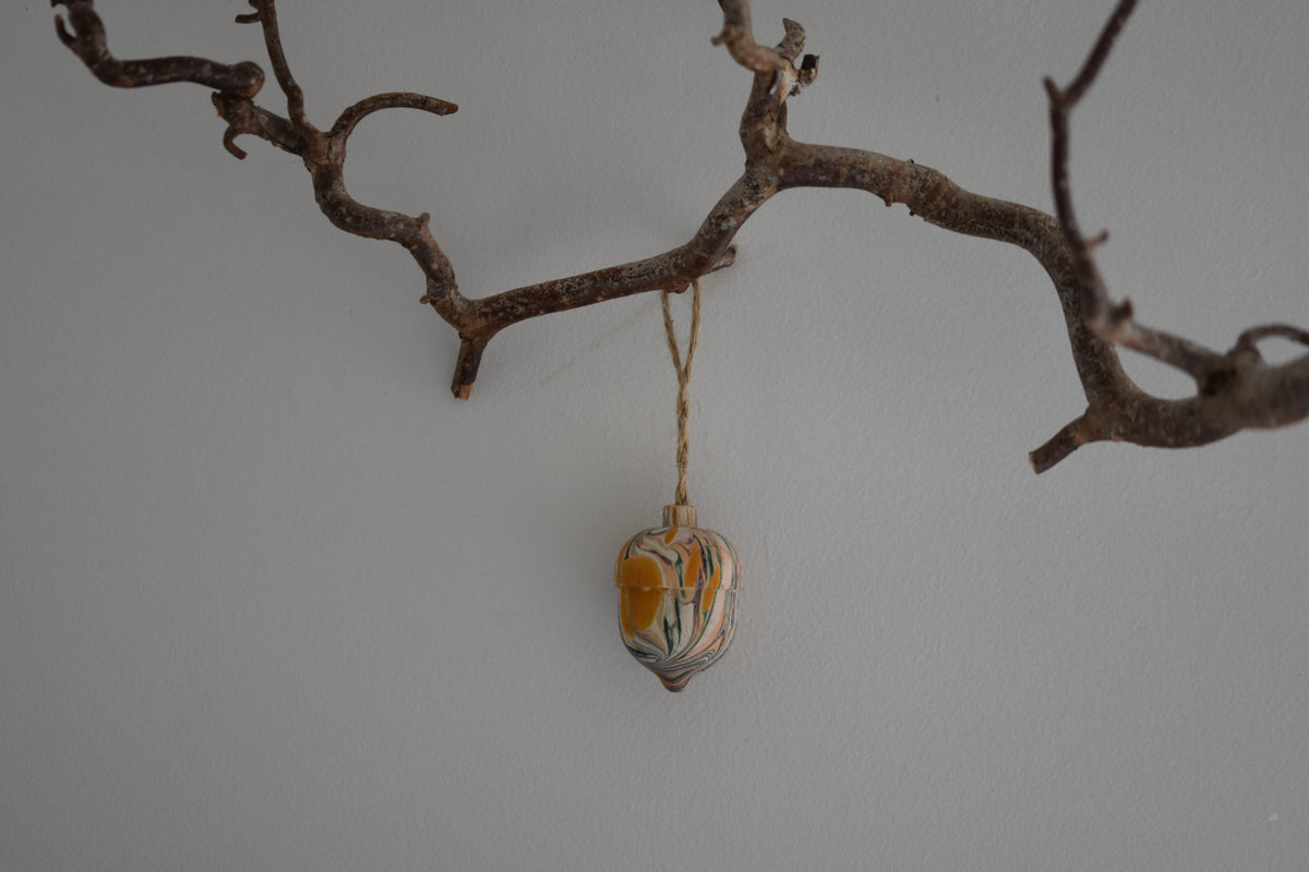 Jemma Lewis colourful marbled wooden acorn decoration Christmas ornament hung from twine on a branch, with a white background.