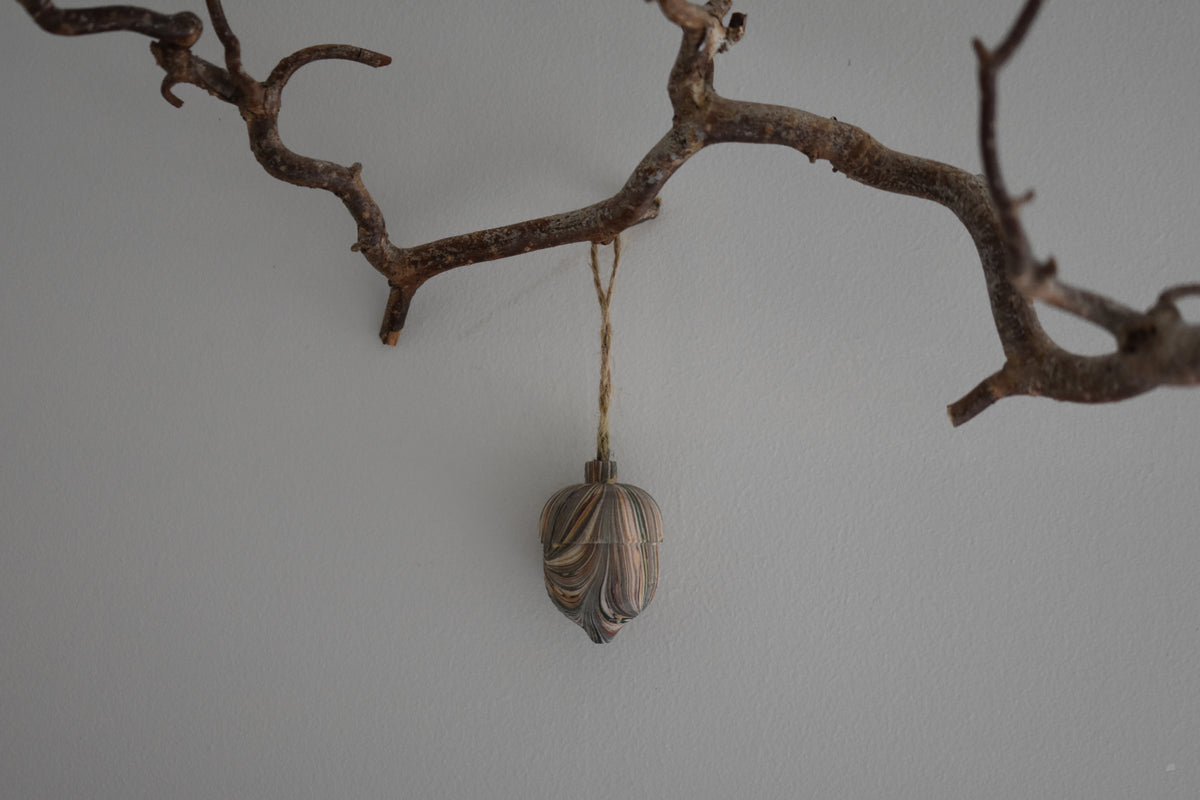 Jemma Lewis colourful marbled wooden acorn decoration Christmas ornament hung from twine on a branch, with a white background.