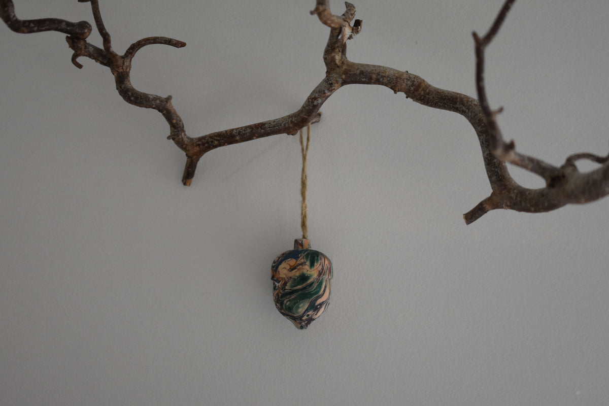 Jemma Lewis colourful marbled wooden acorn decoration Christmas ornament hung from twine on a branch, with a white background.