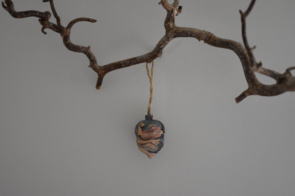 Jemma Lewis colourful marbled wooden acorn decoration Christmas ornament hung from twine on a branch, with a white background.