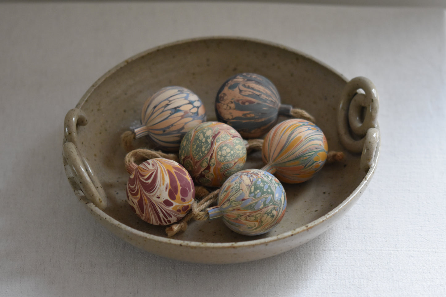 Jemma Lewis colourful marbled wooden round bauble Christmas ornaments with twine, in a ceramic bowl on a beige background.