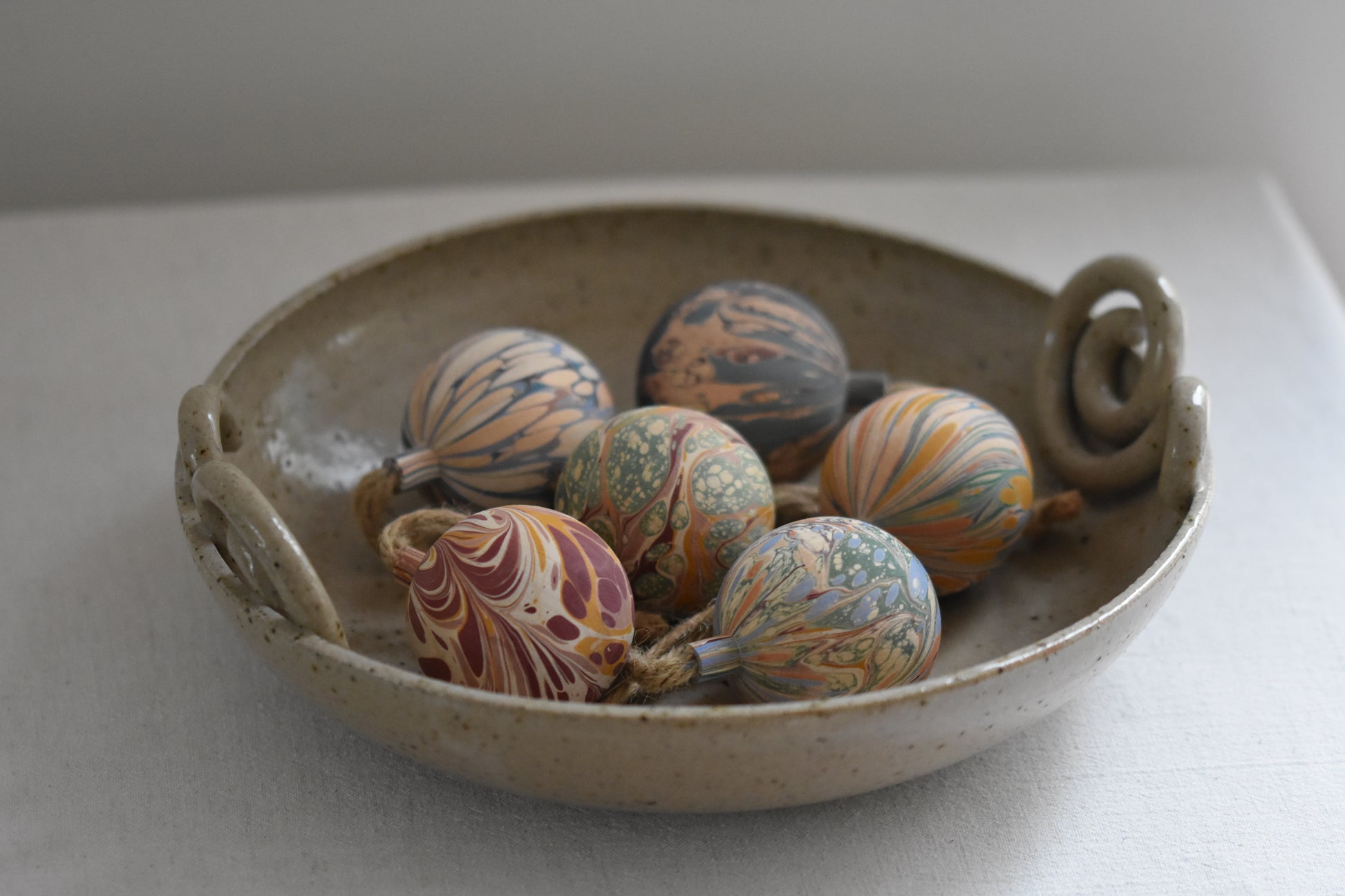 Jemma Lewis colourful marbled wooden round bauble Christmas ornaments with twine, in a ceramic bowl on a beige background.