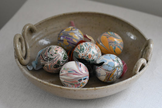 Jemma Lewis colourful marbled round ceramic bauble Christmas ornaments with velvet ribbons, in a ceramic bowl on a beige background.