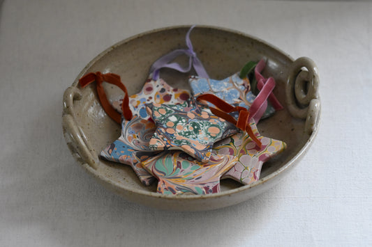 Jemma Lewis marbled ceramic star Christmas ornament decorations with colourful velvet ribbons, in a ceramic bowl on a beige background.