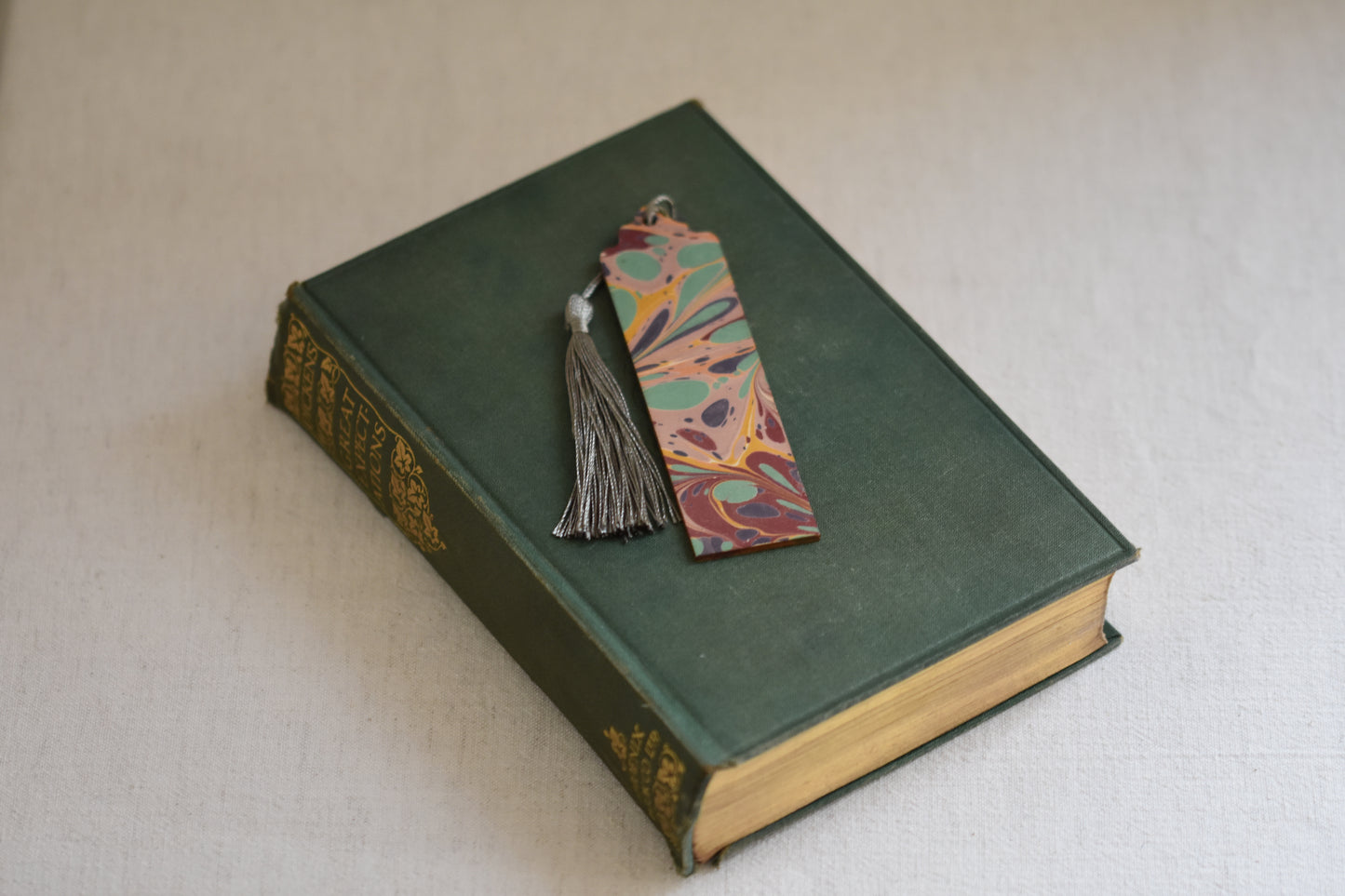 Jemma Lewis colourful marbled wooden bookmark with grey tassel on a vintage green and gold book, with a beige background.