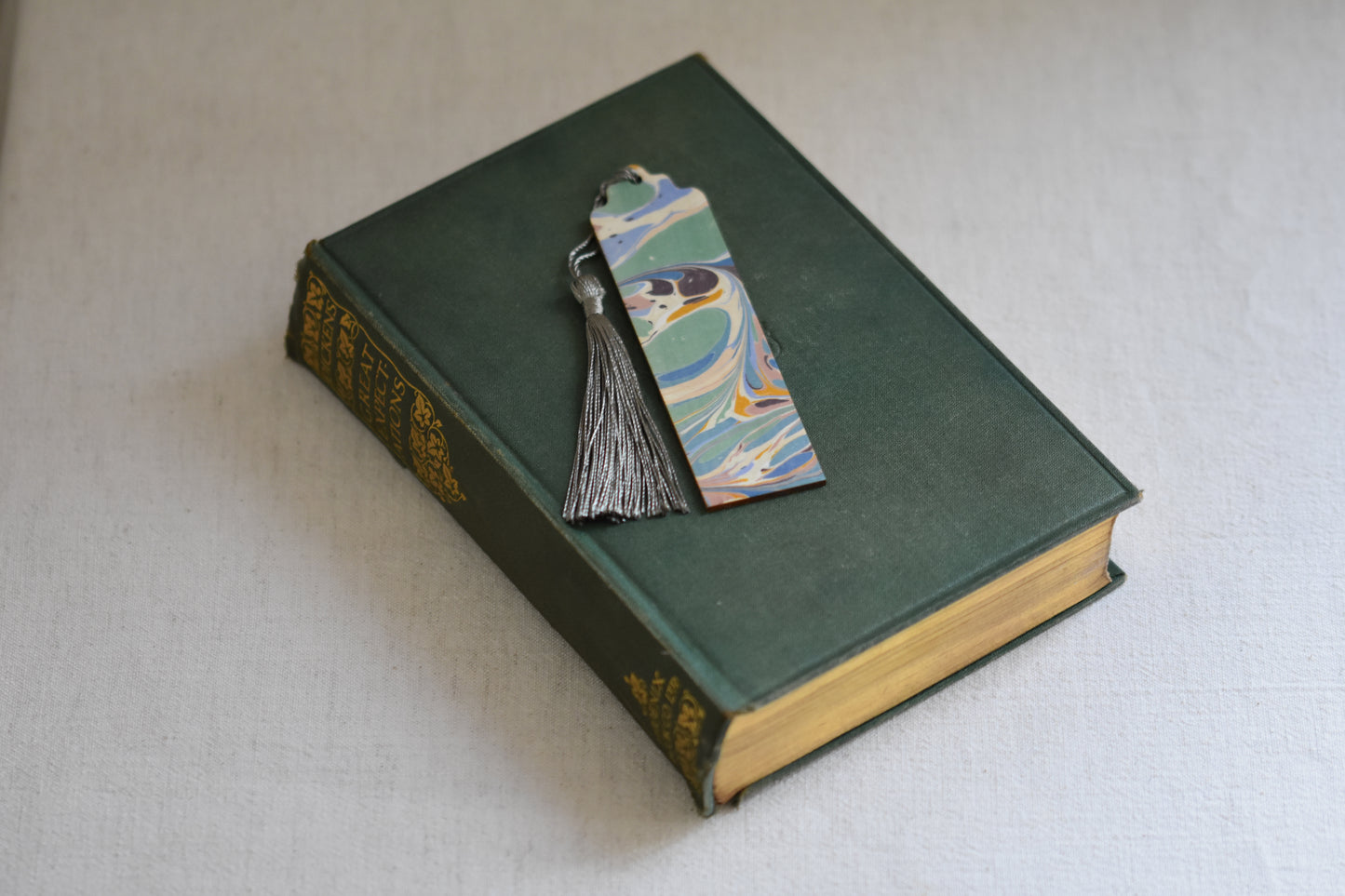 Jemma Lewis colourful marbled wooden bookmark with grey tassel on a vintage green and gold book, with a beige background.